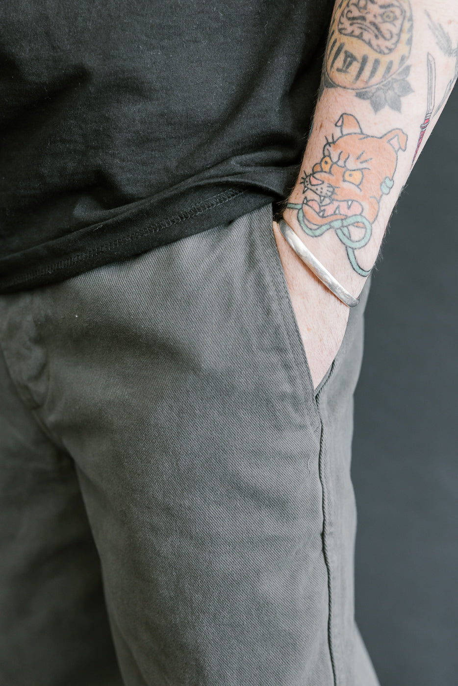 Person wearing a black shirt and gray pants with a tattooed arm against a dark background