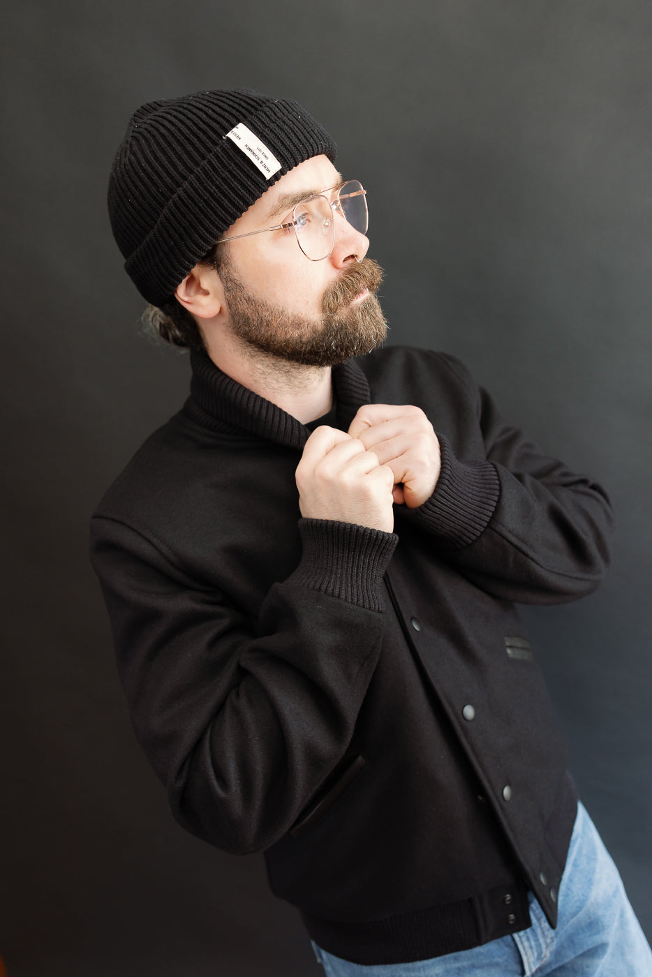 male model wearing an all black varsity jacket with black beanie, washed jeans, 3/4, front view