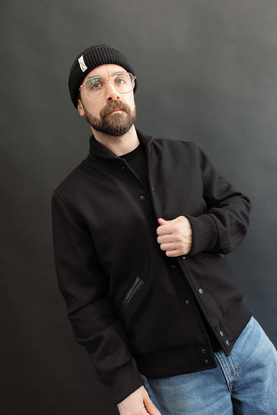 male model wearing an all black varsity jacket with black beanie, washed jeans, holding placket, 3/4 body, front view