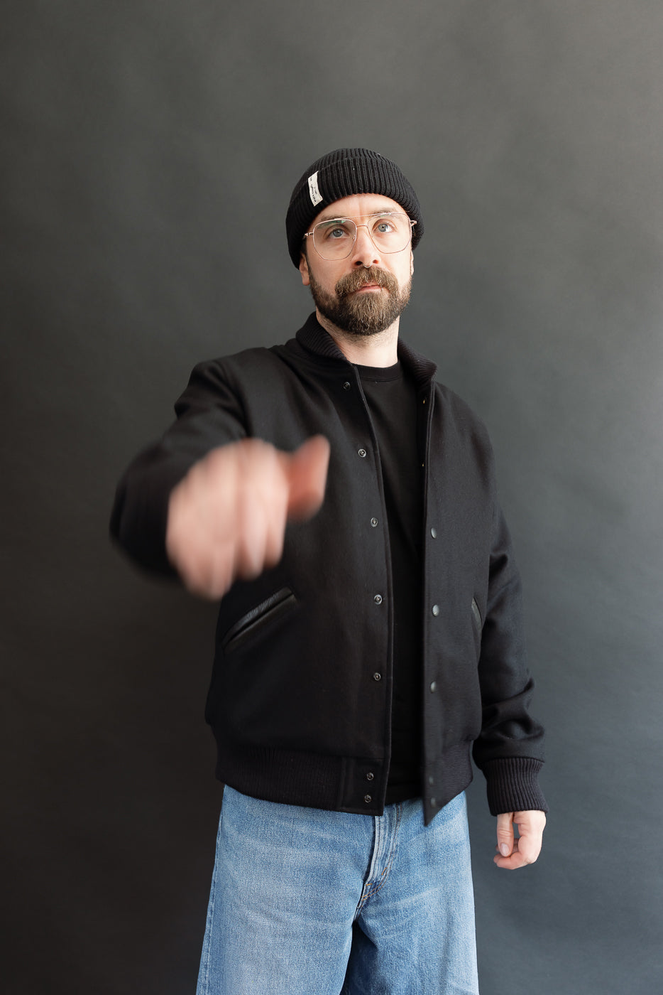 male model wearing an all black varsity jacket with black beanie, washed jeans, opening jacket to show liner, 3/4 body, front view