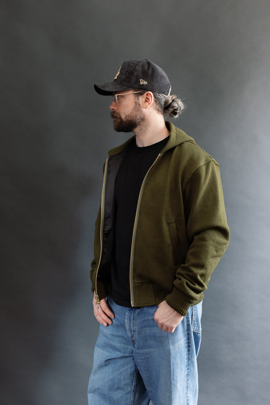 male model wearing an open green knitted zip up hoodie with a black hat, t-shirt, and washed jeans, 3/4 body, side view