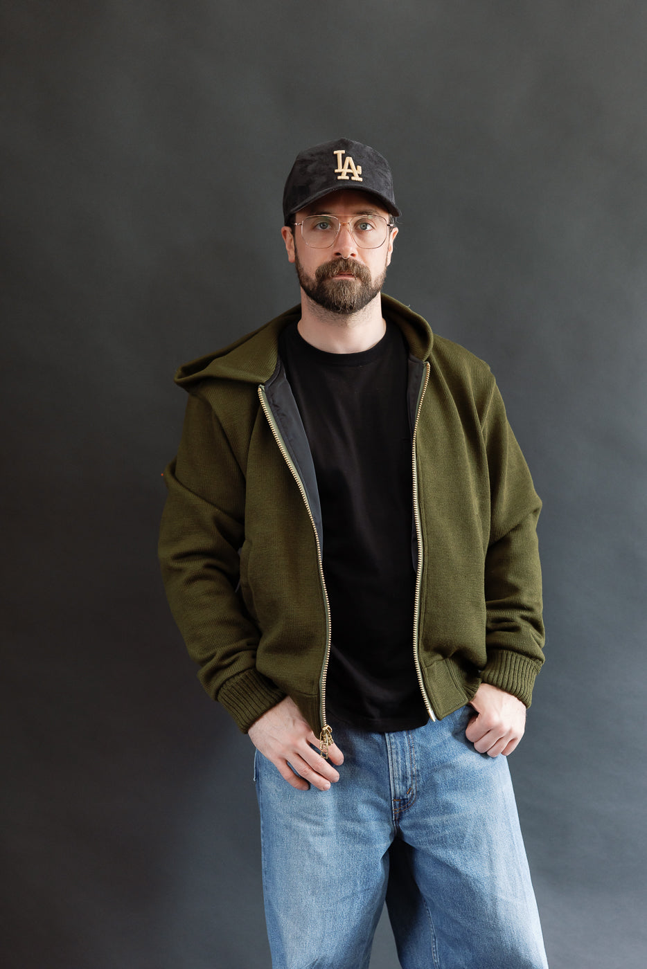 male model wearing an open green knitted zip up hoodie with a black hat, t-shirt, and washed jeans, 3/4 body, front view