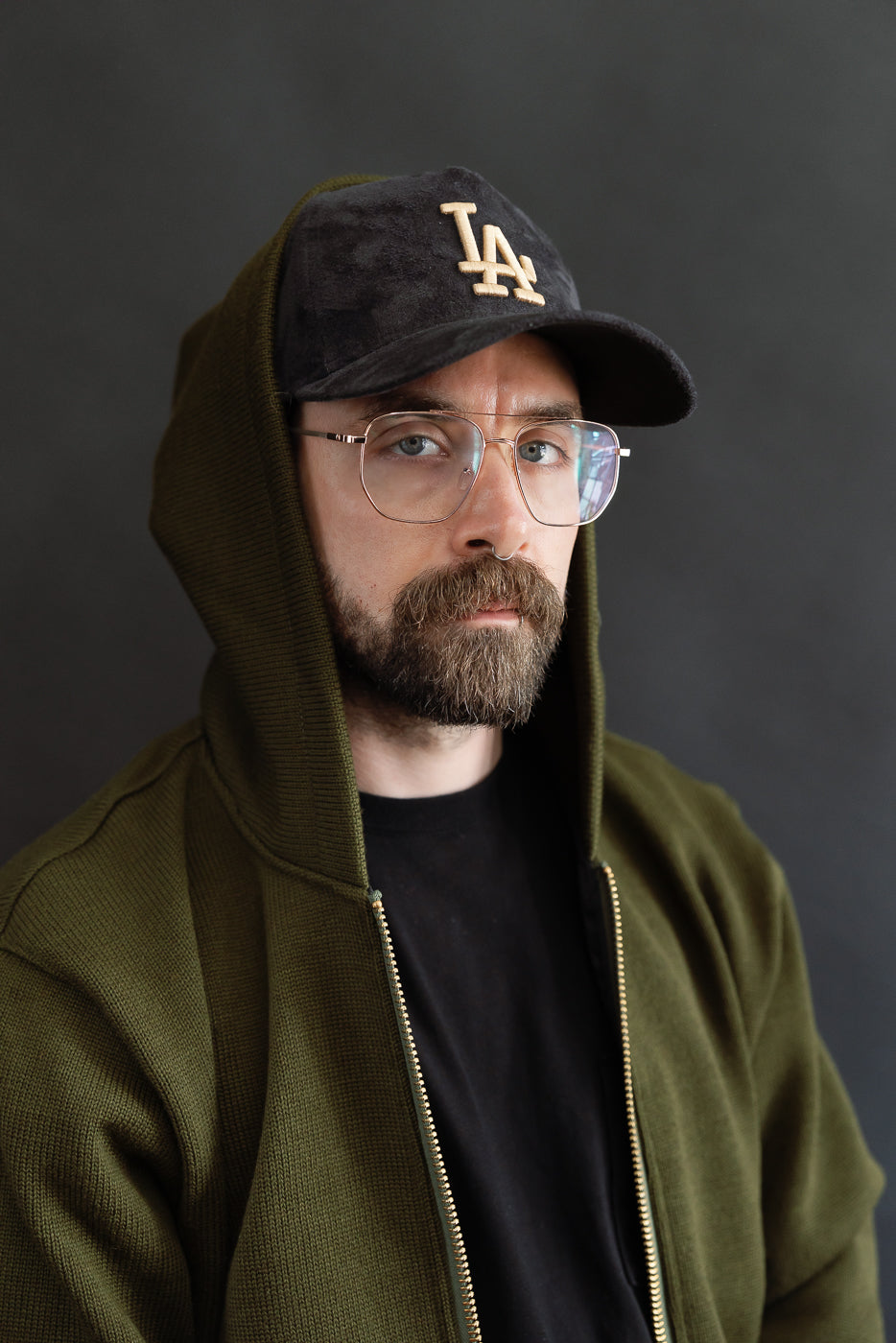close up of male model wearing a black LA cap and black t-shirt with olive green knitted hoodie layered over the top, hood up over hat