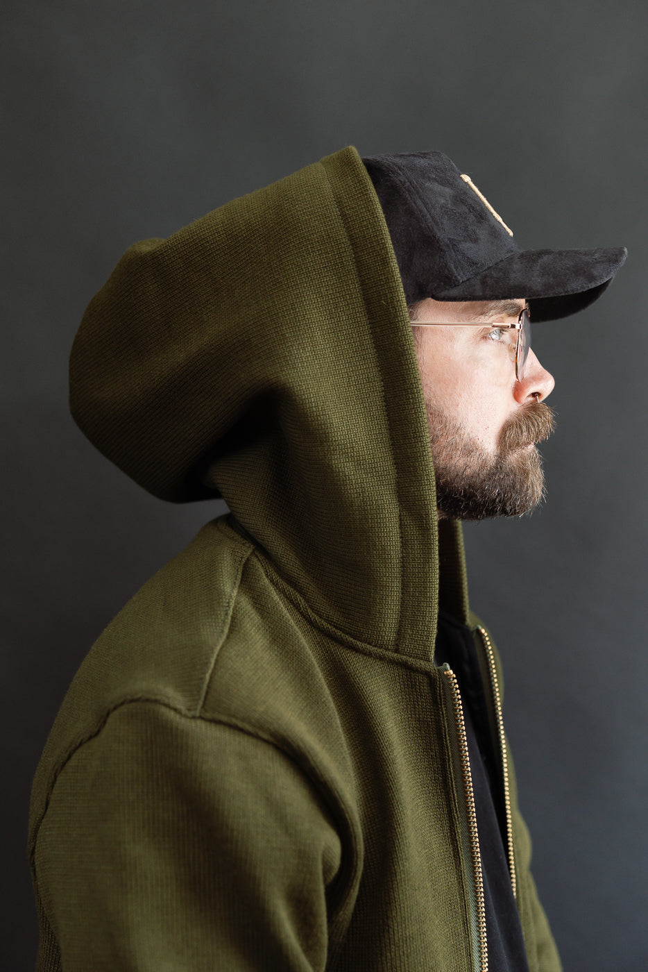 close up of male model wearing a black LA cap and black t-shirt with olive green knitted hoodie layered over the top, hood up over hat, side view
