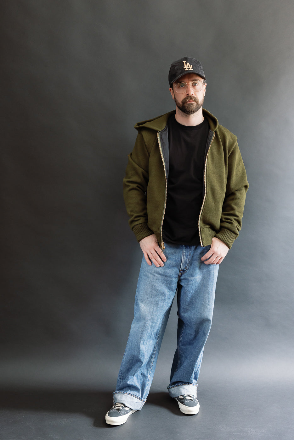 male model wearing an open green knitted zip up hoodie with a black hat, t-shirt, and washed jeans, full body, front view