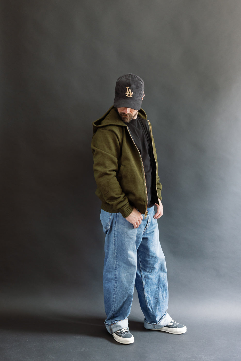 male model wearing an open green knitted zip up hoodie with a black hat, t-shirt, and washed jeans, full body, side view