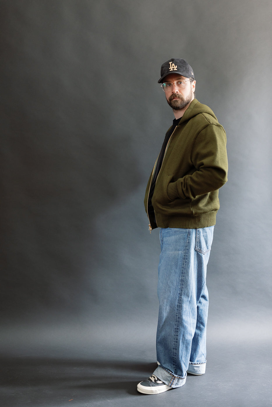 male model wearing an open green knitted zip up hoodie with a black hat, t-shirt, and washed jeans, full body, side view