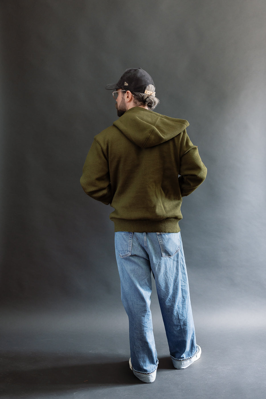 male model wearing an open green knitted zip up hoodie with a black hat, t-shirt, and washed jeans, full body, back view