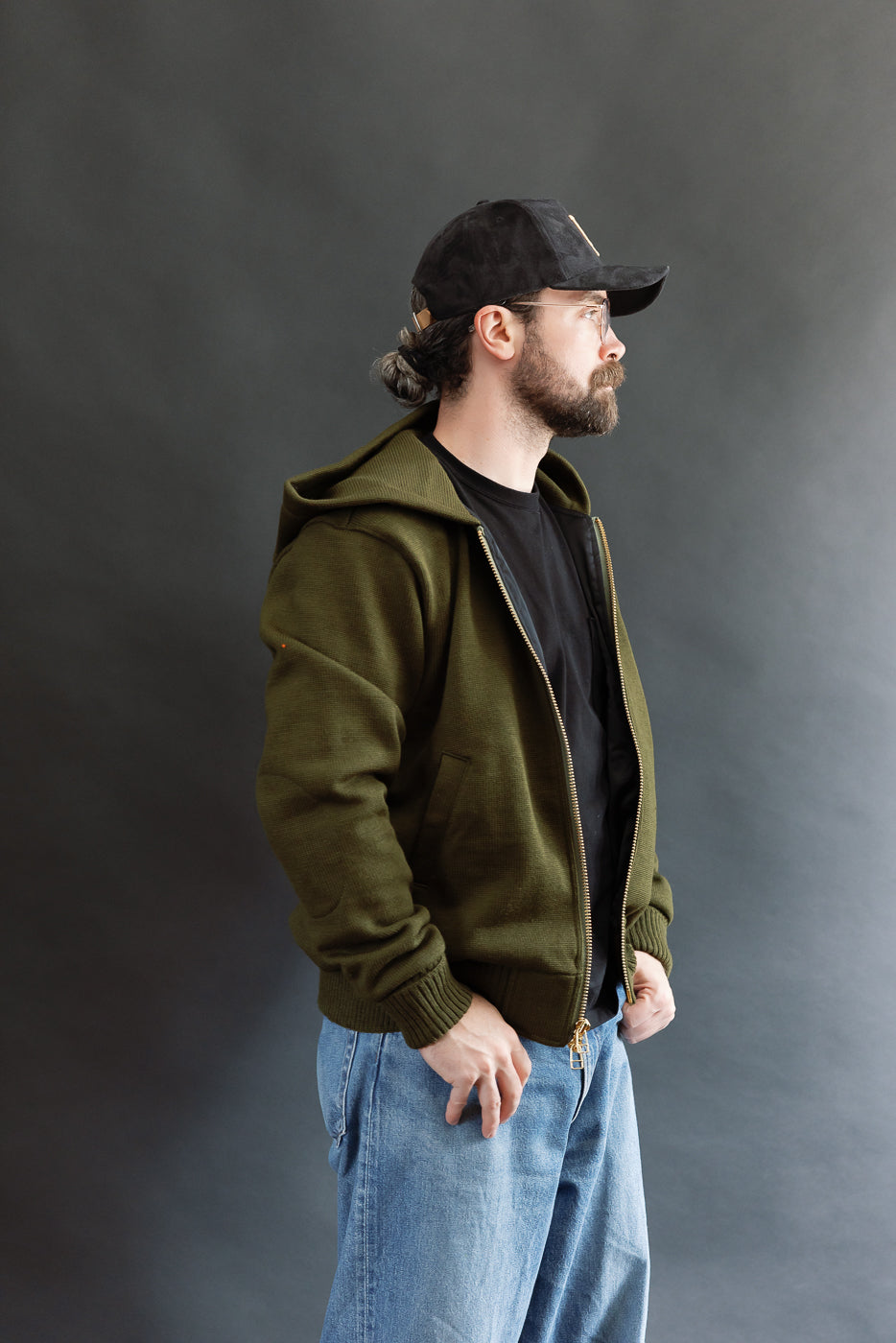 male model wearing an open green knitted zip up hoodie with a black hat, t-shirt, and washed jeans, 3/4 body, side view