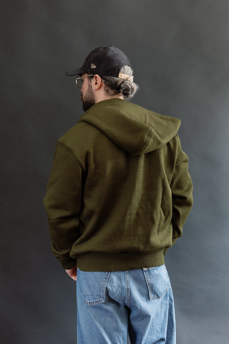male model wearing an open green knitted zip up hoodie with a black hat, t-shirt, and washed jeans, 3/4 body, back view