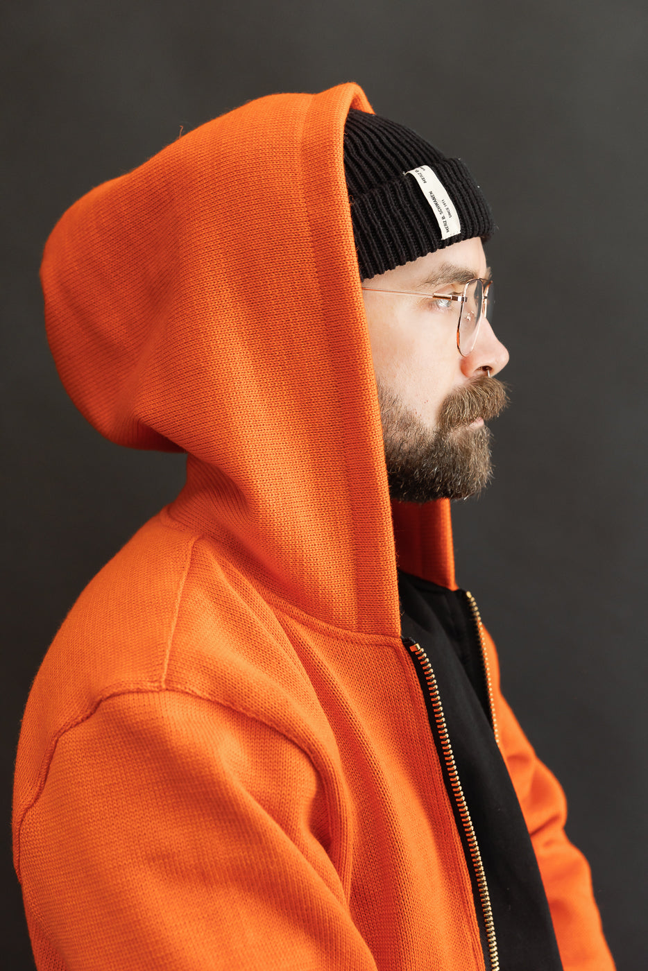 Male model wearing a knitted orange hoodie (open), black beanie, black shirt, close up, side view, hood up