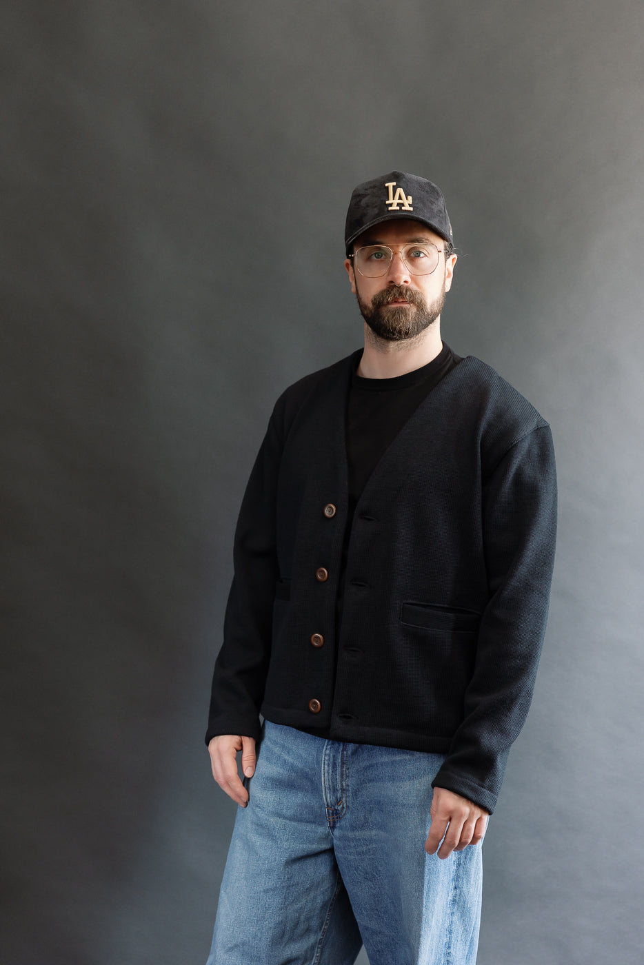 Male model wearing Black Dant slouchy cardigan with brown buttons and washed baggy blue jeans and black cap, 3/4 front view