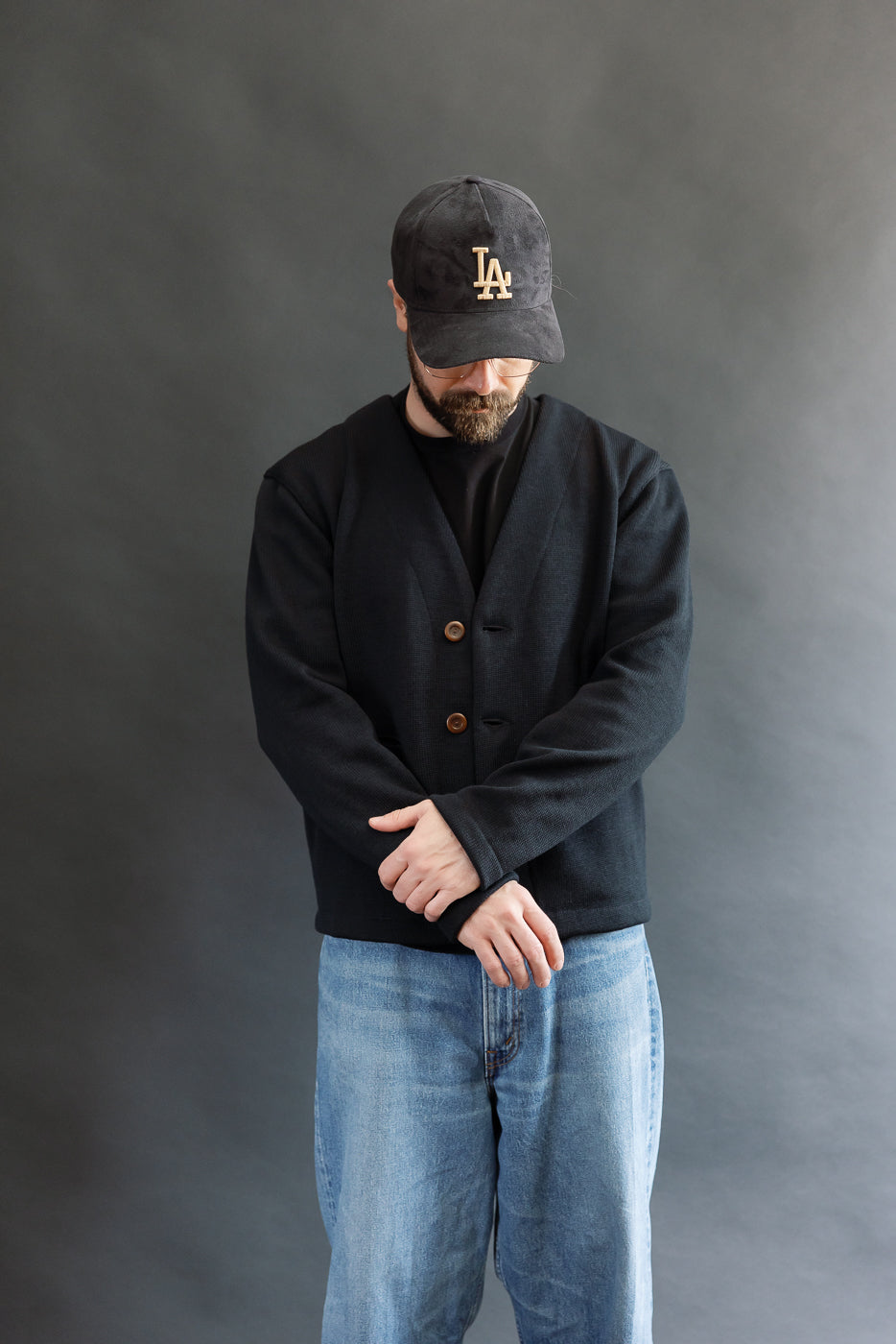 Male model wearing Black Dant slouchy cardigan with brown buttons and washed baggy blue jeans and black cap, 3/4 front view arms crossed looking down