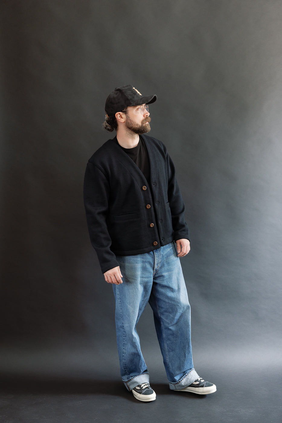 Male model wearing Black Dant slouchy cardigan with brown buttons and washed baggy blue jeans and black cap, full body, side view