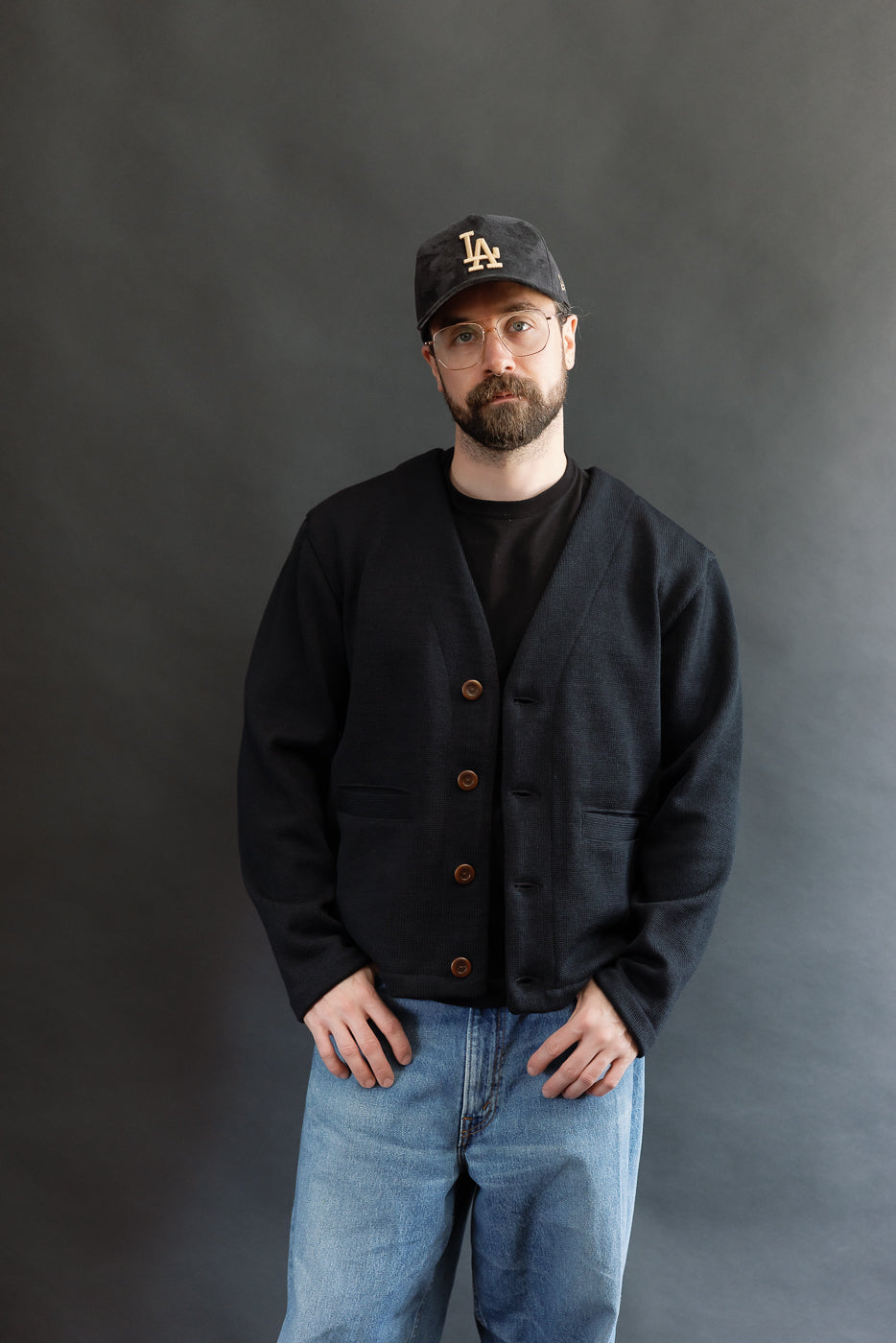 Male model wearing Black Dant slouchy cardigan with brown buttons and washed baggy blue jeans and black cap, 3/4 view