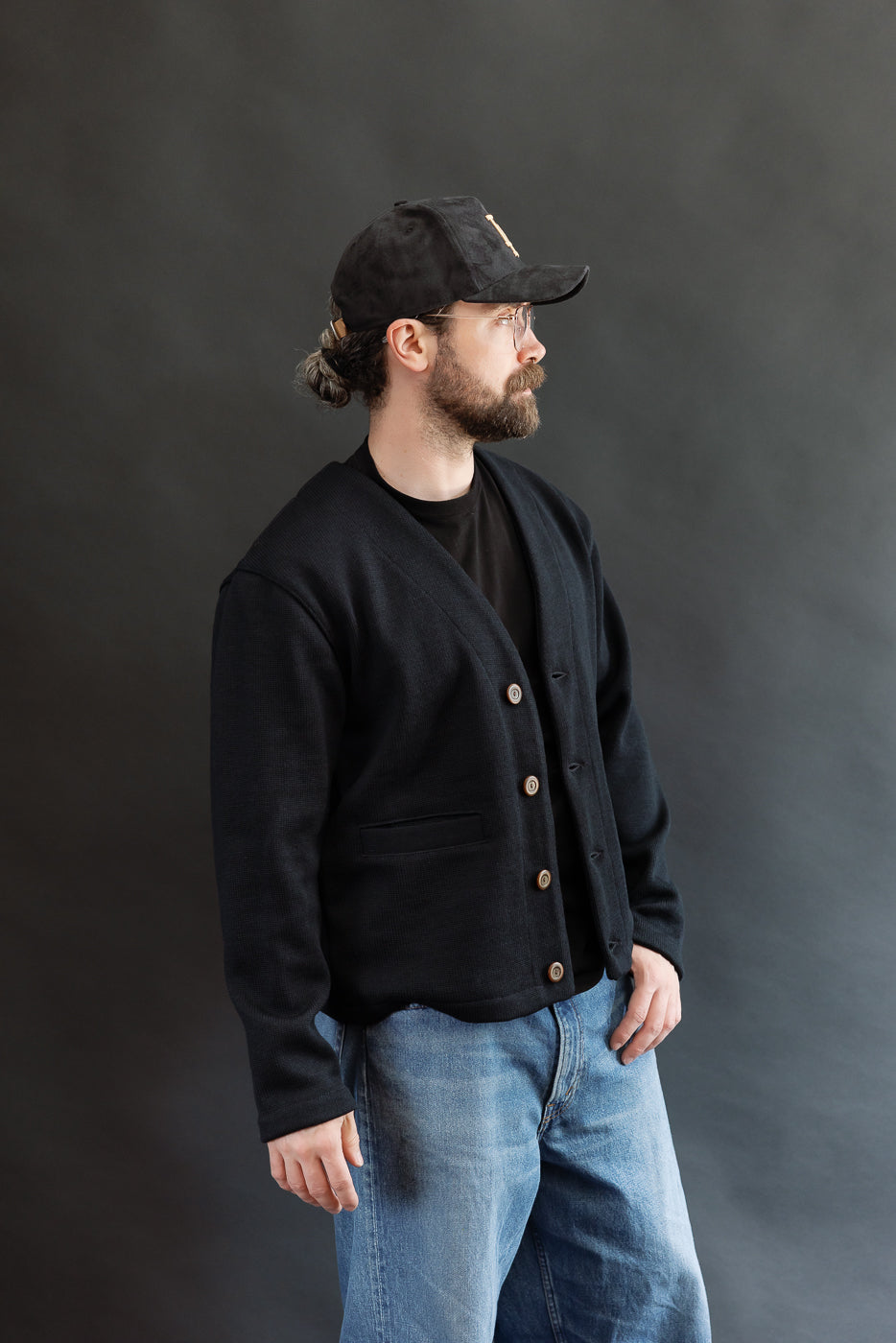 Male model wearing Black Dant slouchy cardigan with brown buttons and washed baggy blue jeans and black cap, 3/4 side view