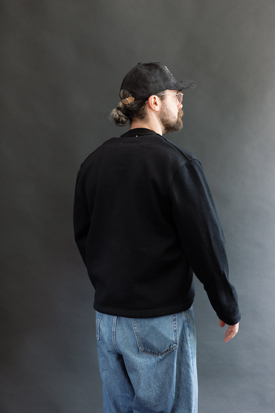 Male model wearing Black Dant slouchy cardigan with brown buttons and washed baggy blue jeans and black cap, 3/4 back view