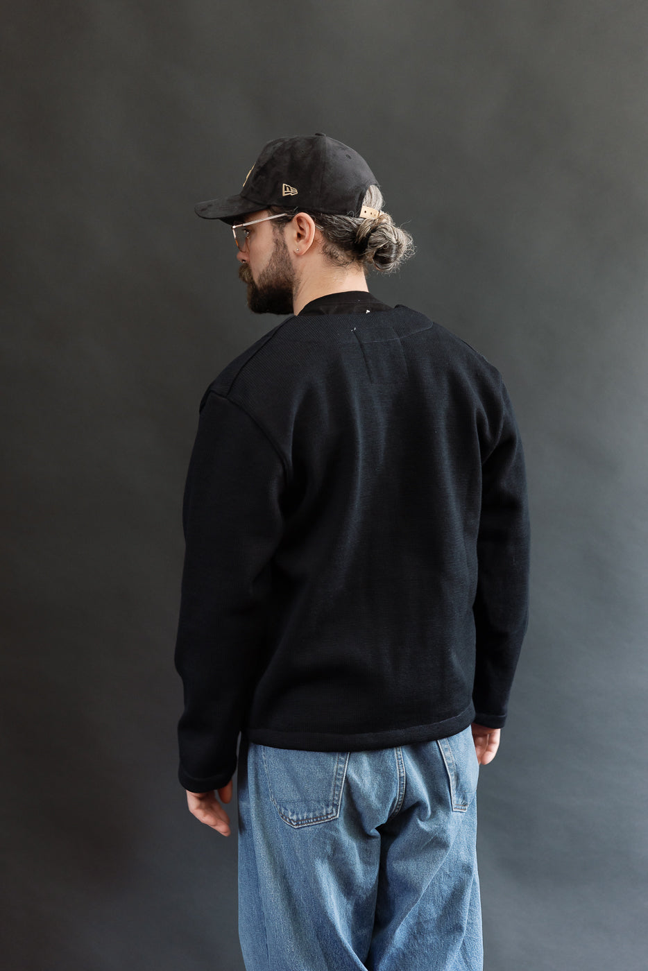Male model wearing Black Dant slouchy cardigan with brown buttons and washed baggy blue jeans and black cap, 3/4 back view