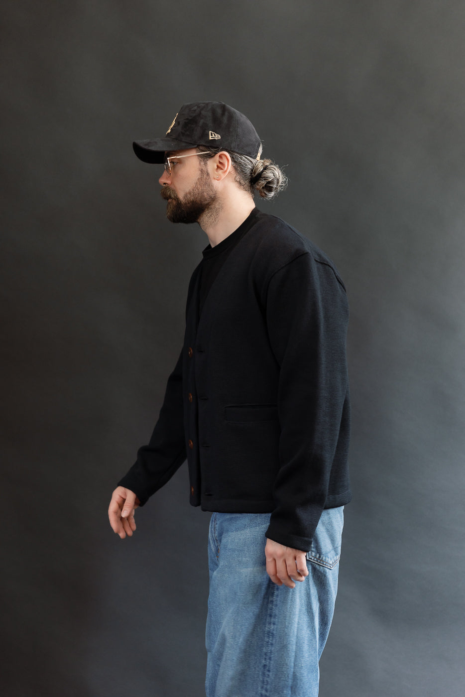 Male model wearing Black Dant slouchy cardigan with brown buttons and washed baggy blue jeans and black cap, 3/4 side view