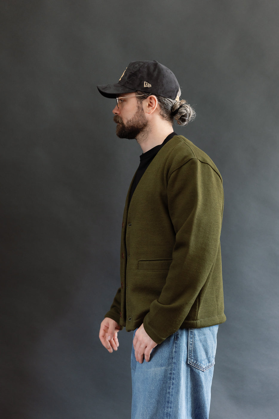 Male model wearing an olive green slouchy cardigan w black t-shirt, black cap, 3/4 length, side view