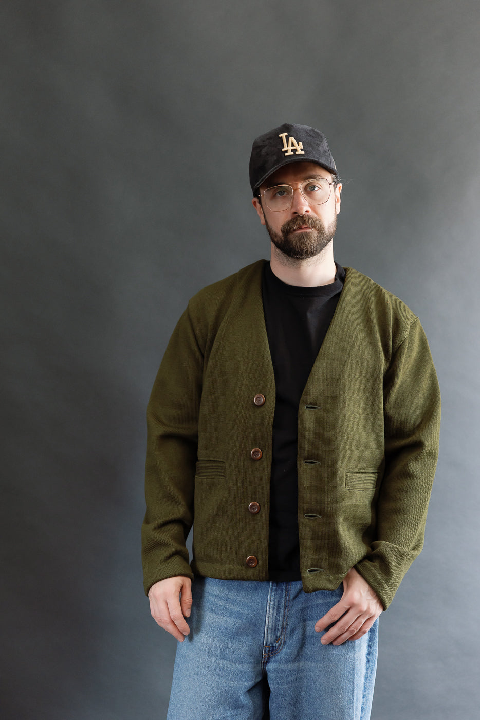 Male model wearing an olive green slouchy cardigan w black t-shirt, black cap, 3/4 length, front view