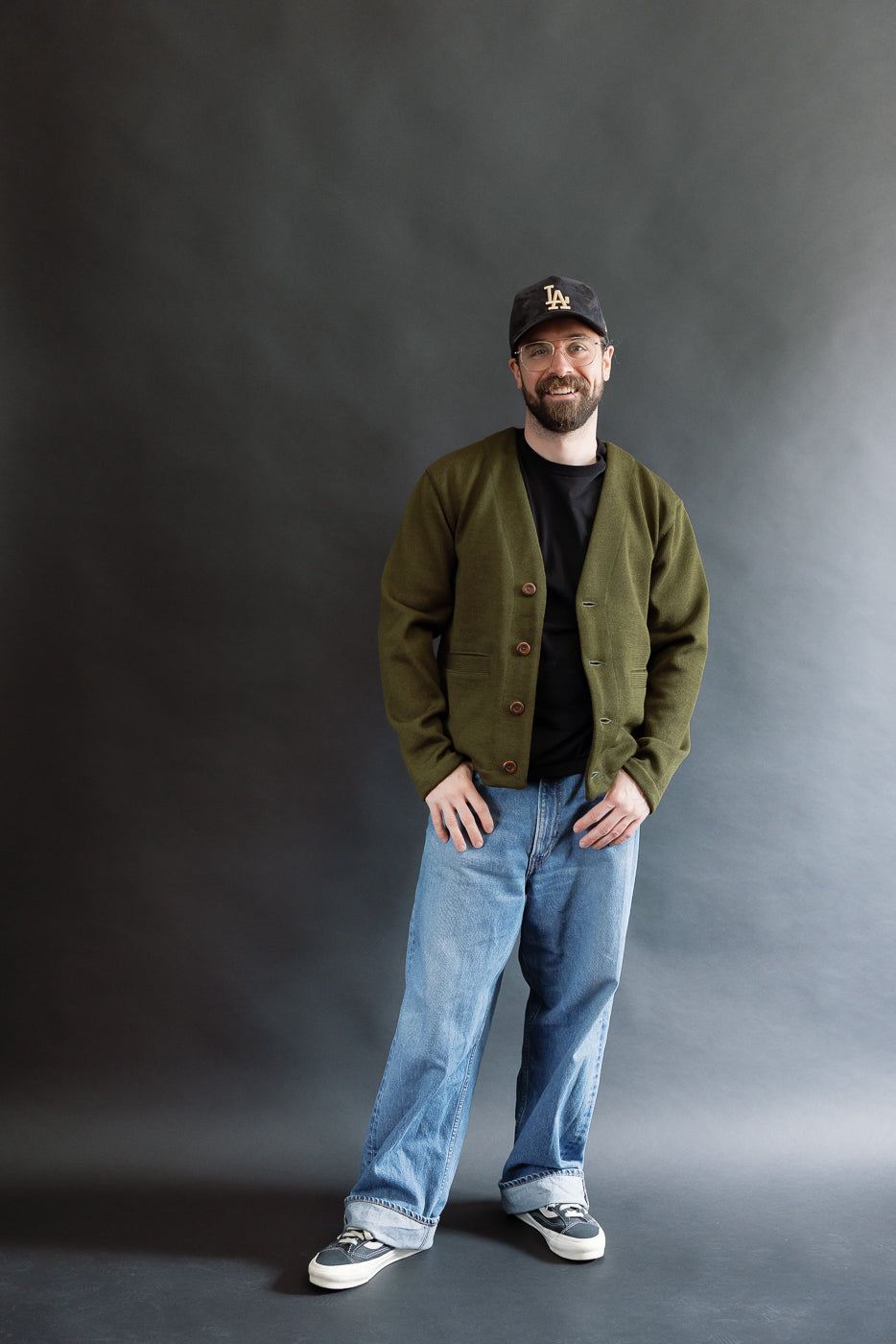 Male model wearing an olive green slouchy cardigan, black cap, and baggy washed blue jeans against a black background, full body, front view