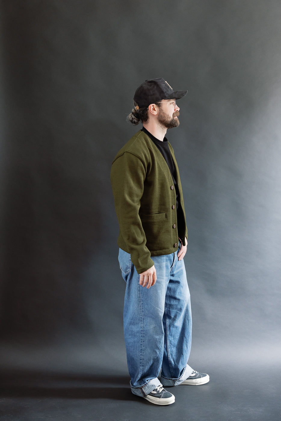 Male model wearing an olive green slouchy cardigan, black cap, and baggy washed blue jeans against a black background, full body, side view