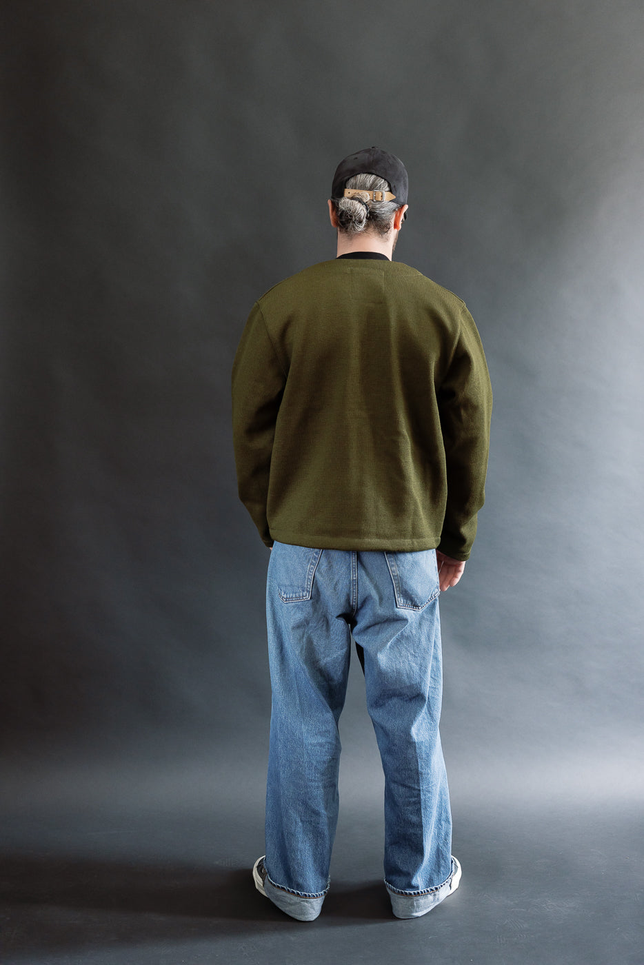 Male model wearing an olive green slouchy cardigan, black cap, and baggy washed blue jeans against a black background, full body, back view