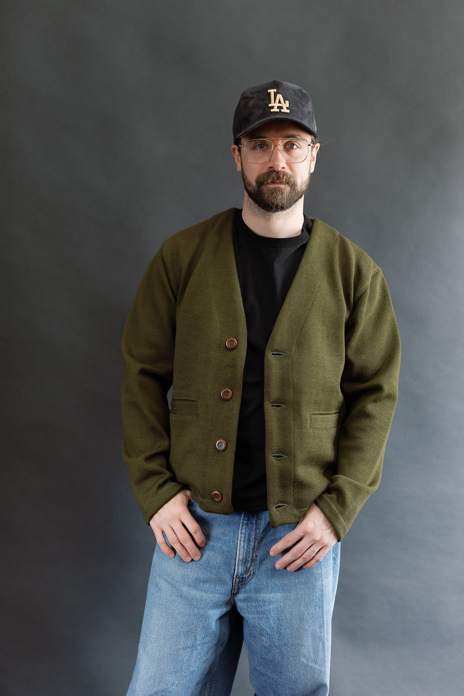 Male model wearing an olive green slouchy cardigan w black t-shirt, black cap, 3/4 length, front view