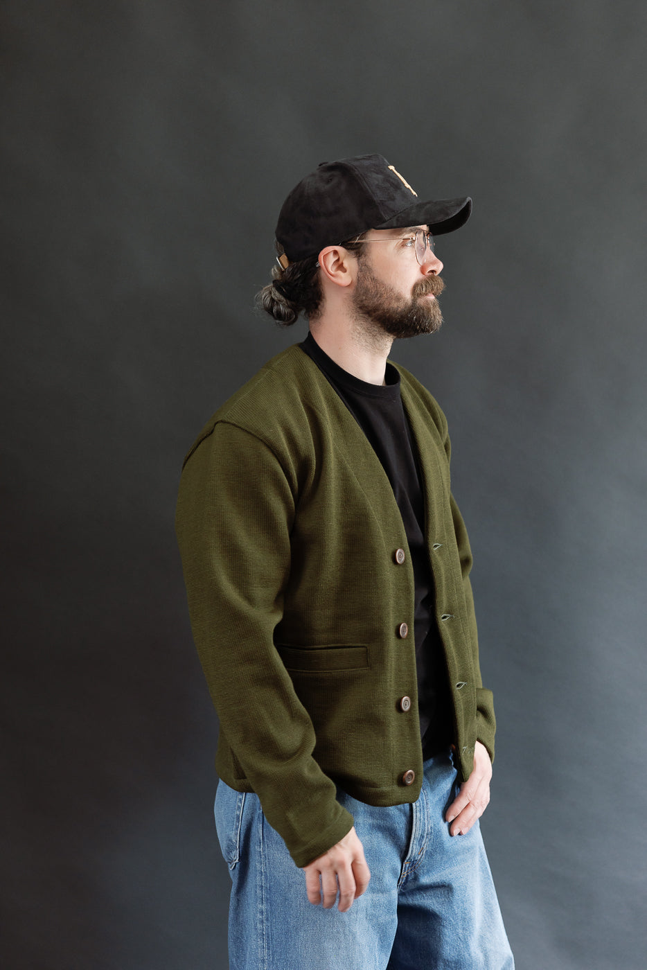 Male model wearing an olive green slouchy cardigan w black t-shirt, black cap, 3/4 length, side view