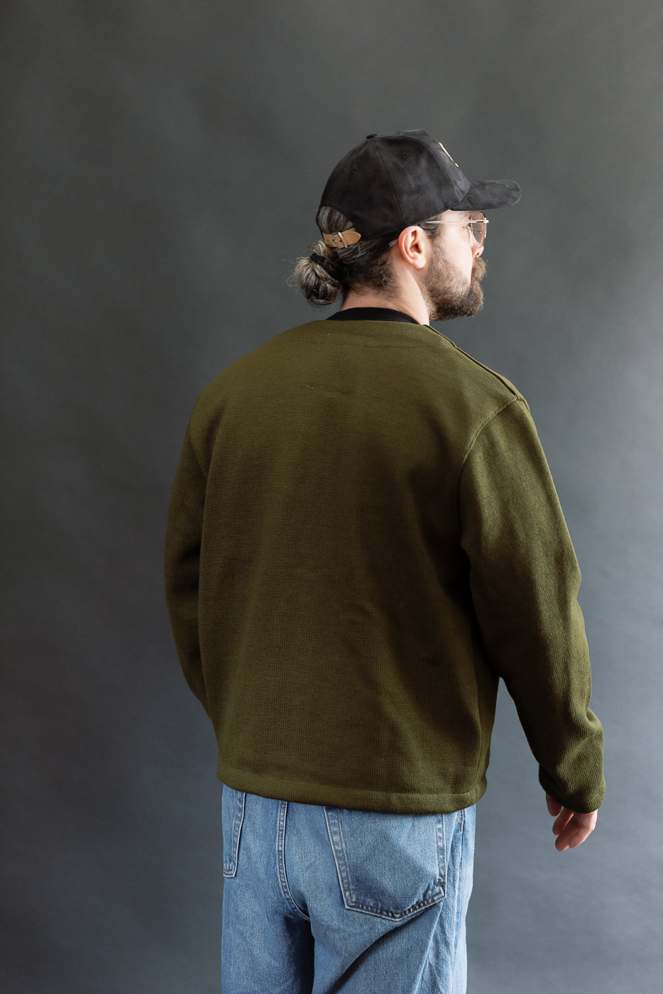 Male model wearing an olive green slouchy cardigan w black t-shirt, black cap, 3/4 length, back view
