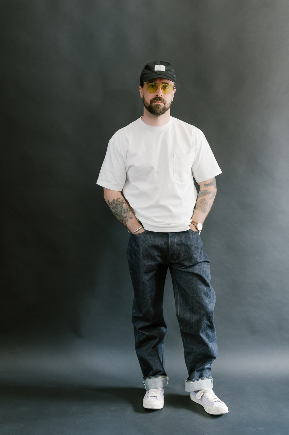 Man wearing a white t-shirt, dark jeans, and a black cap against a gray background