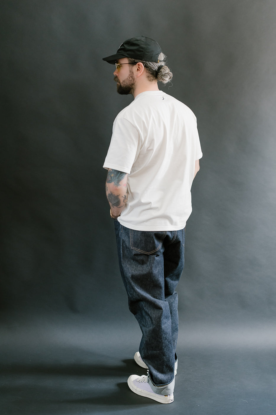 Man wearing a white t-shirt, dark jeans, and a black cap against a gray background