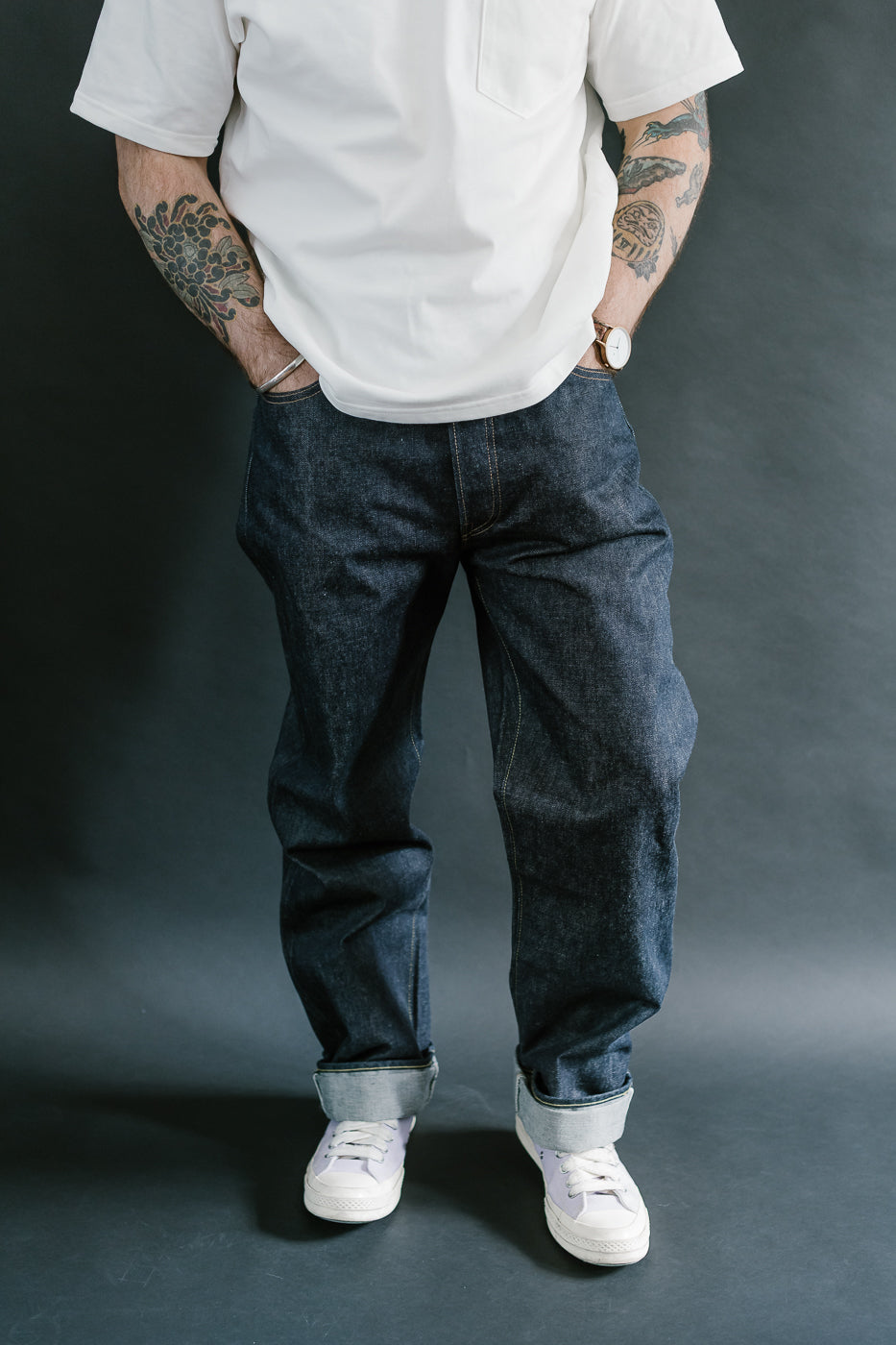 Person wearing a white shirt, dark blue jeans, and white sneakers against a gray background