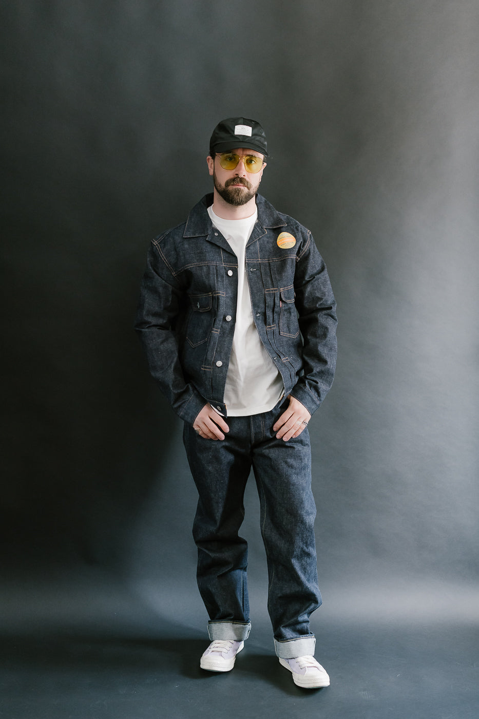 Man wearing a denim jacket, white shirt, jeans, and sunglasses against a dark background