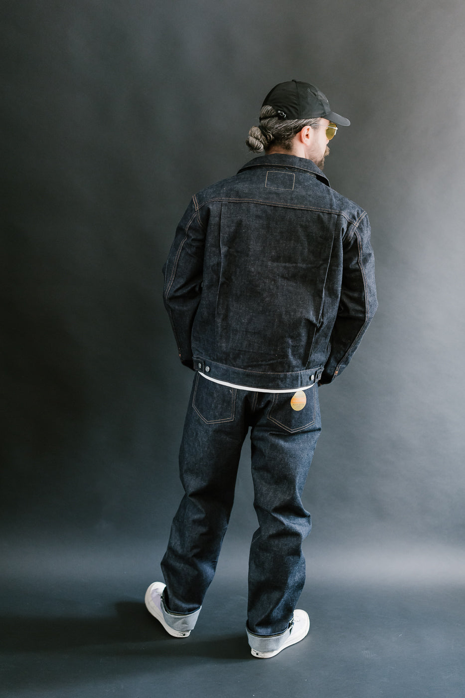 Person wearing a dark denim jacket and jeans standing against a gray background