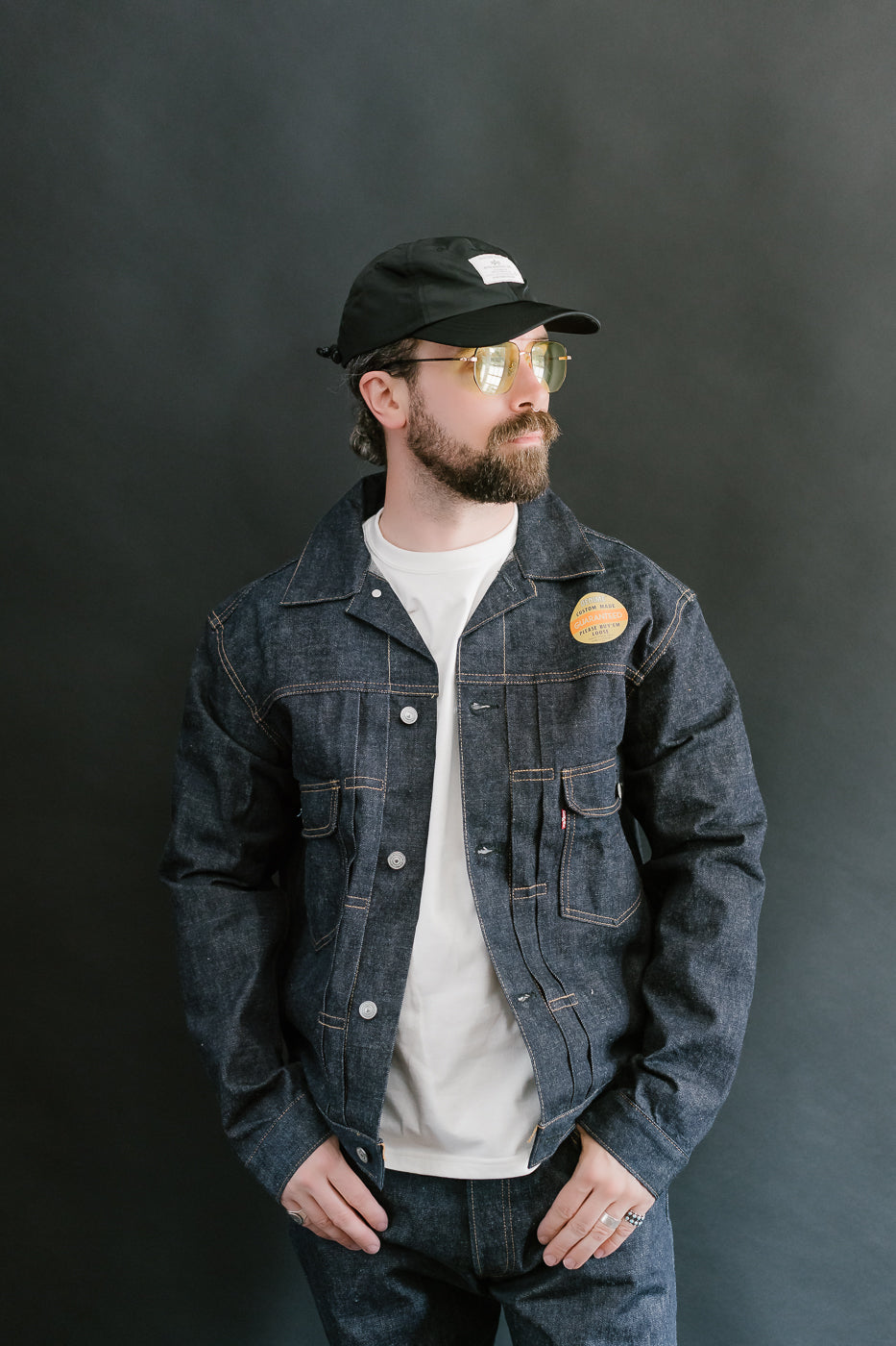 Man wearing a denim jacket, white t-shirt, and black cap against a dark background