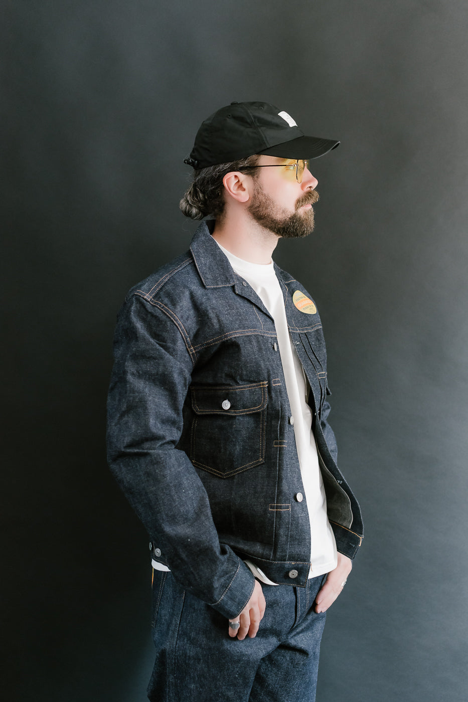 Man wearing a denim jacket, white shirt, and black cap against a dark background