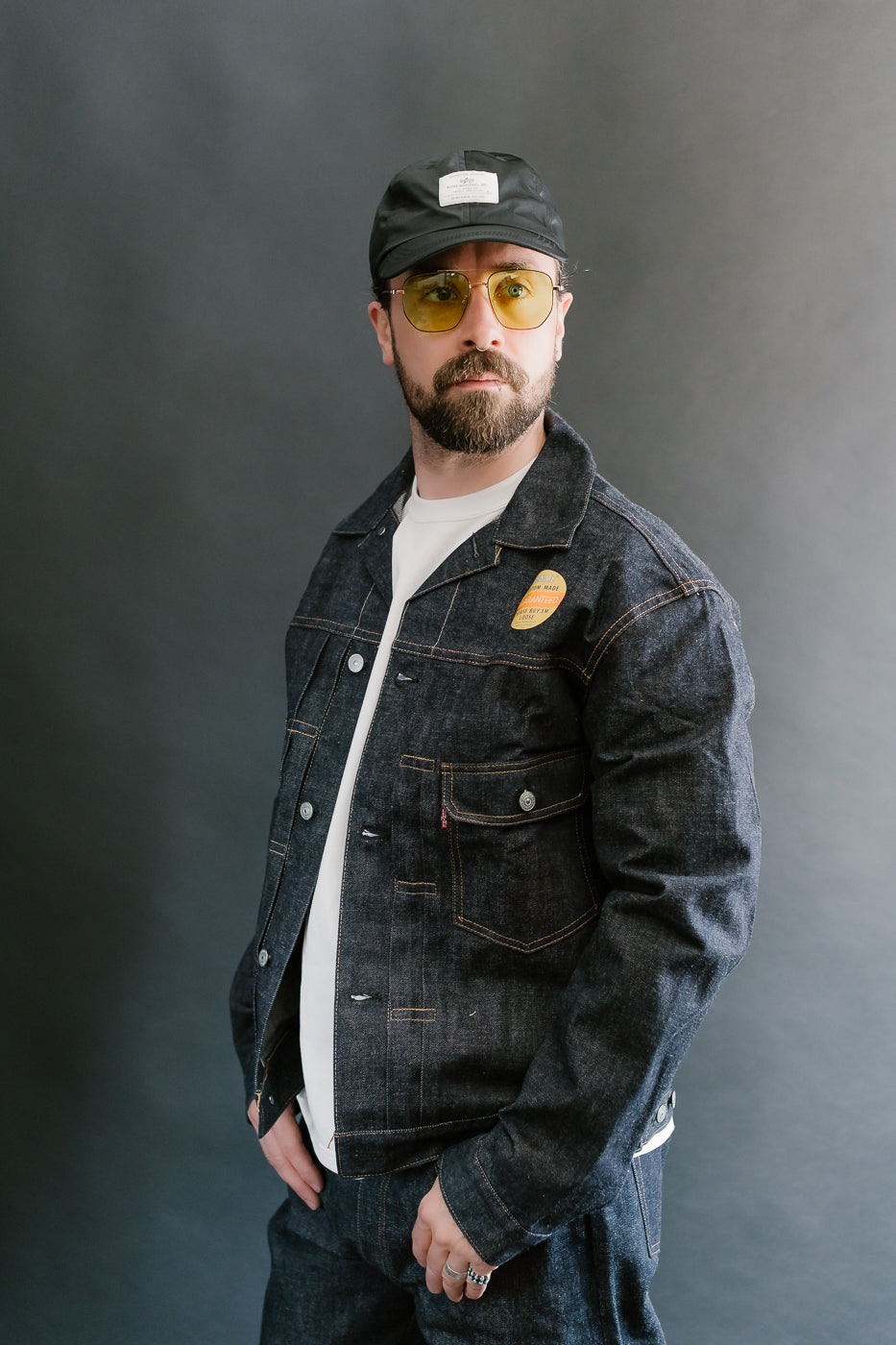 Man wearing a denim jacket, cap, and sunglasses against a gray background