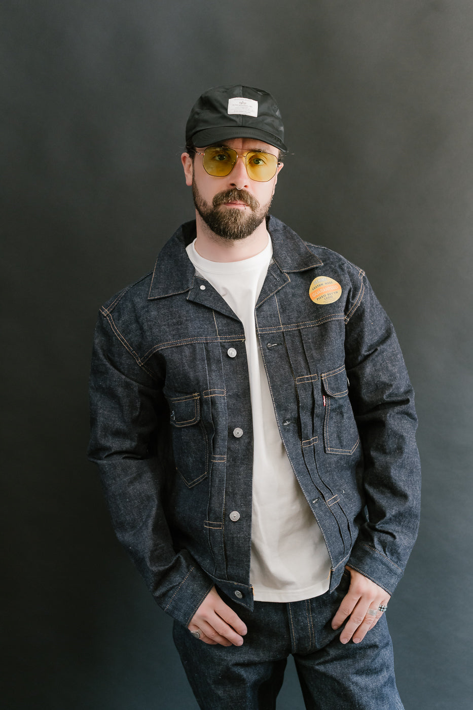 Man wearing a denim jacket, white shirt, and black cap against a dark background