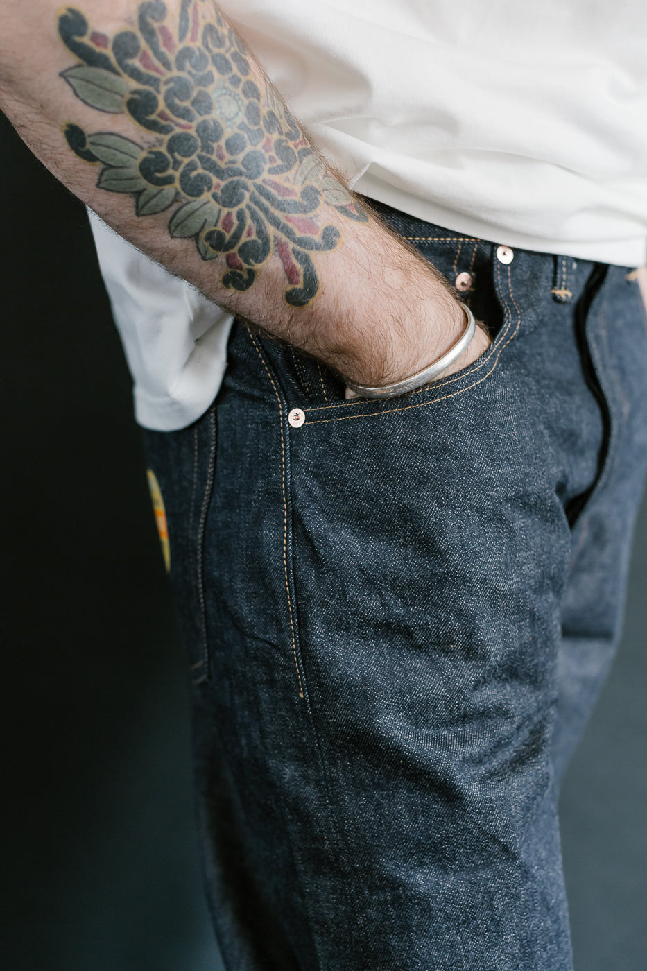 Person wearing blue jeans with a tattoo on their arm, against a dark background