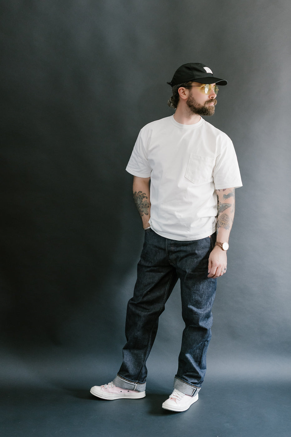 Man wearing a white t-shirt, dark jeans, and a black cap against a gray background