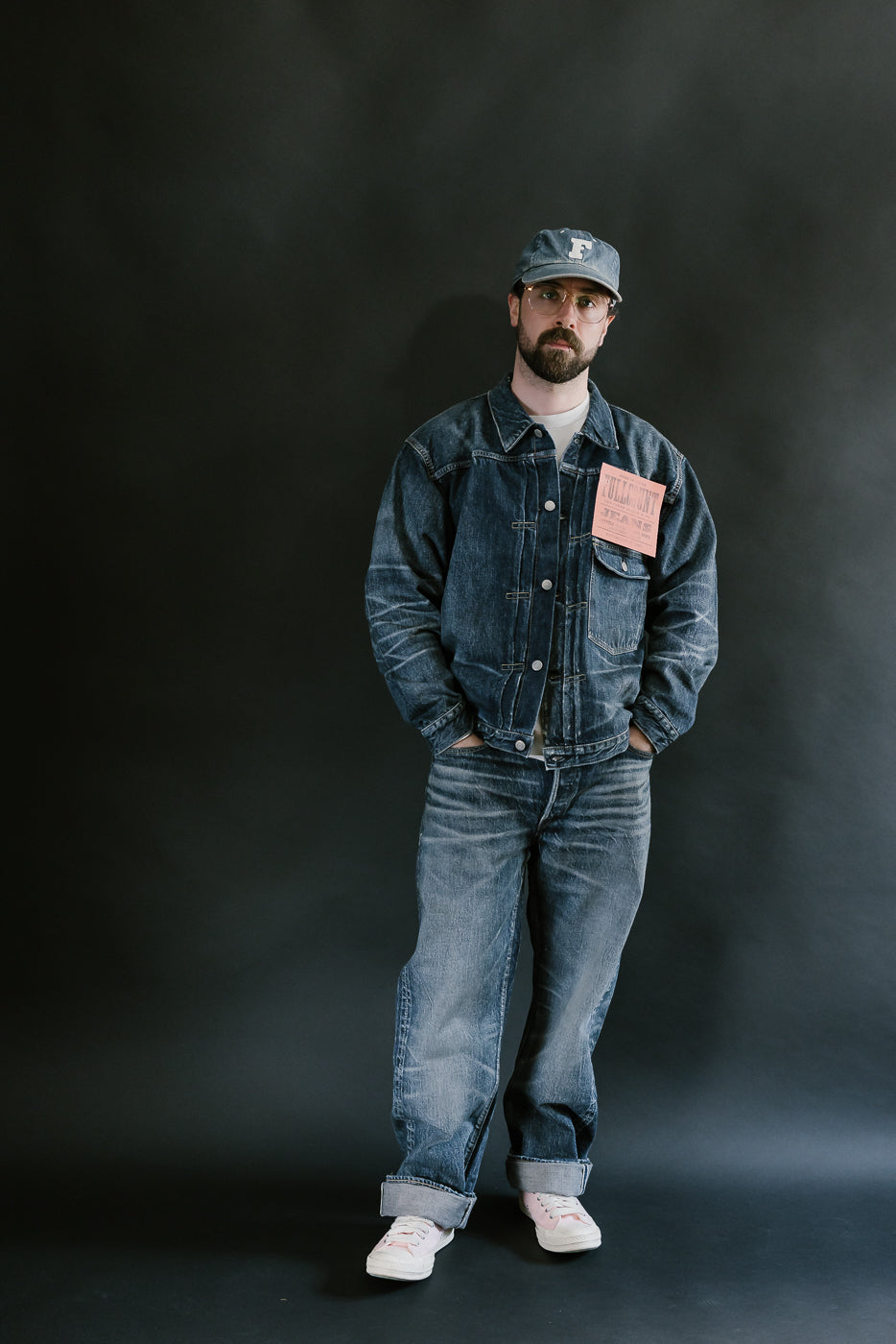 Bearded man wearing heavily washed denim jacket and jeans, pink shoes, washed denim cap, and pink shoes with a plain black background, full body, front view.