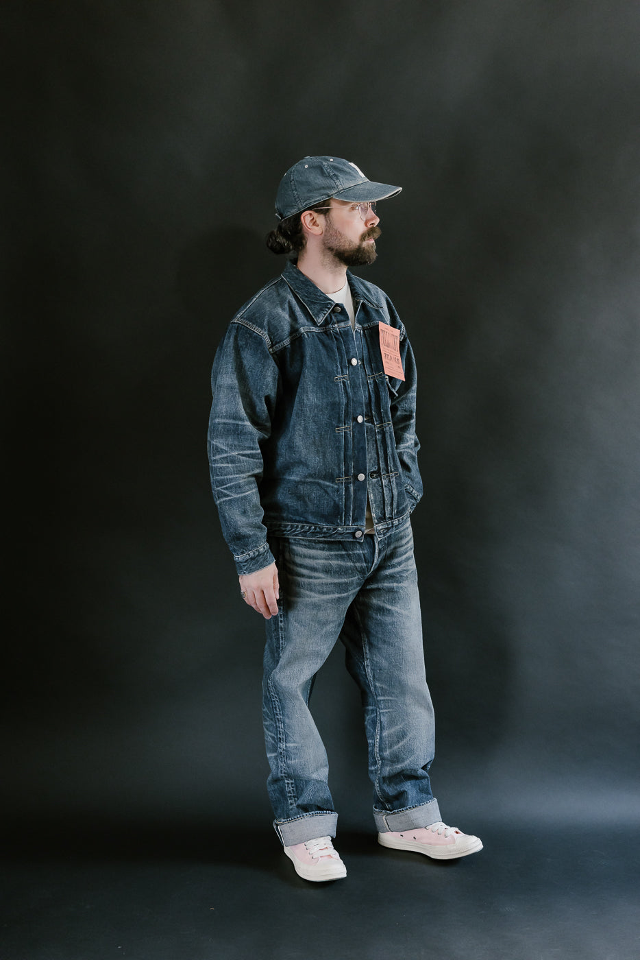 Bearded man wearing heavily washed denim jacket and jeans, pink shoes, washed denim cap, and pink shoes with a plain black background, full body, side view.