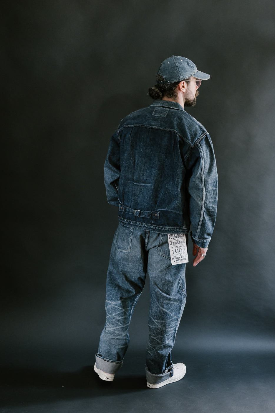 Bearded man wearing heavily washed denim jacket and jeans, pink shoes, washed denim cap, and pink shoes with a plain black background, full body, back view.