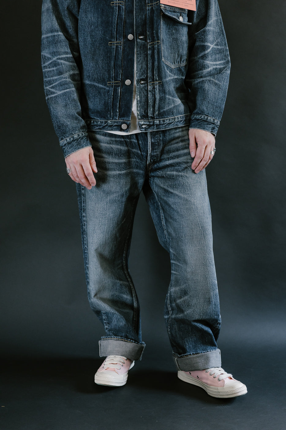 Man wearing heavily washed denim jacket and jeans,and pink shoes with a plain black background, 3/4 body, front view.