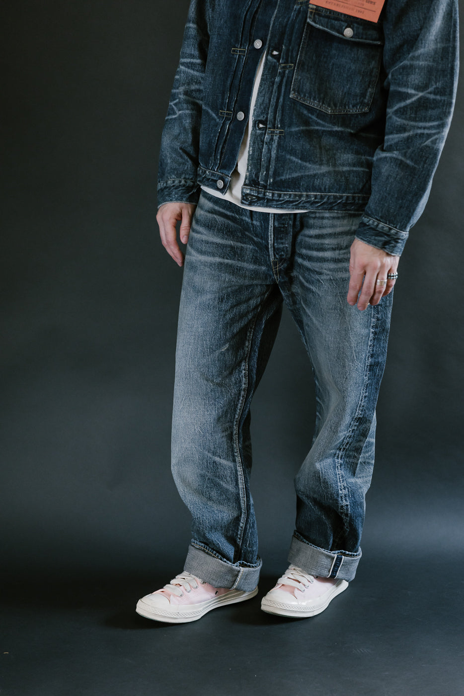 Man wearing heavily washed denim jacket and jeans,and pink shoes with a plain black background, 3/4 body, side view.