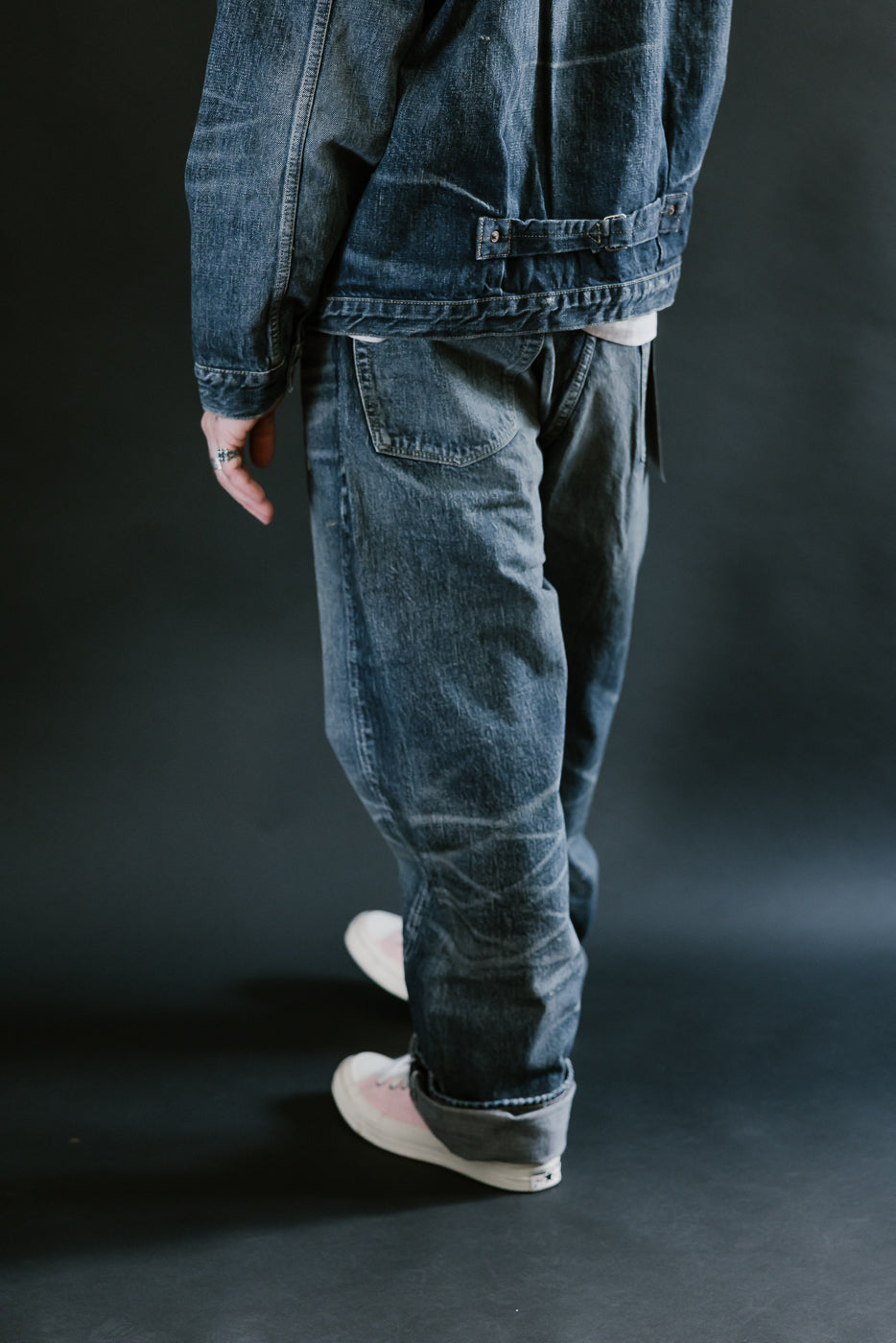Man wearing heavily washed denim jacket and jeans,and pink shoes with a plain black background, 3/4 body, back view.