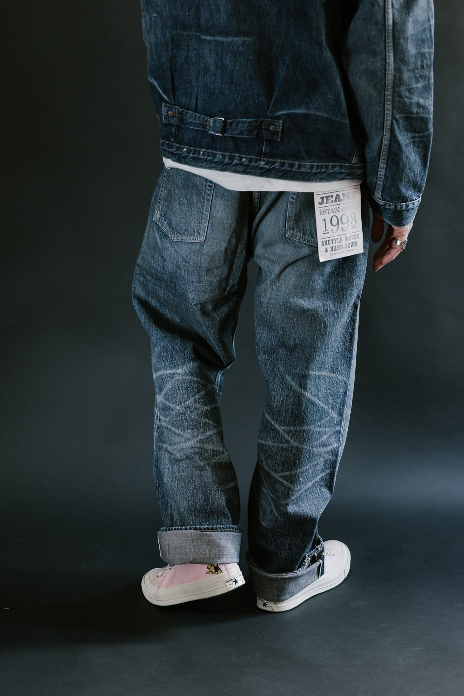 Man wearing heavily washed denim jacket and jeans,and pink shoes with a plain black background, 3/4 body, back view, leg raised.