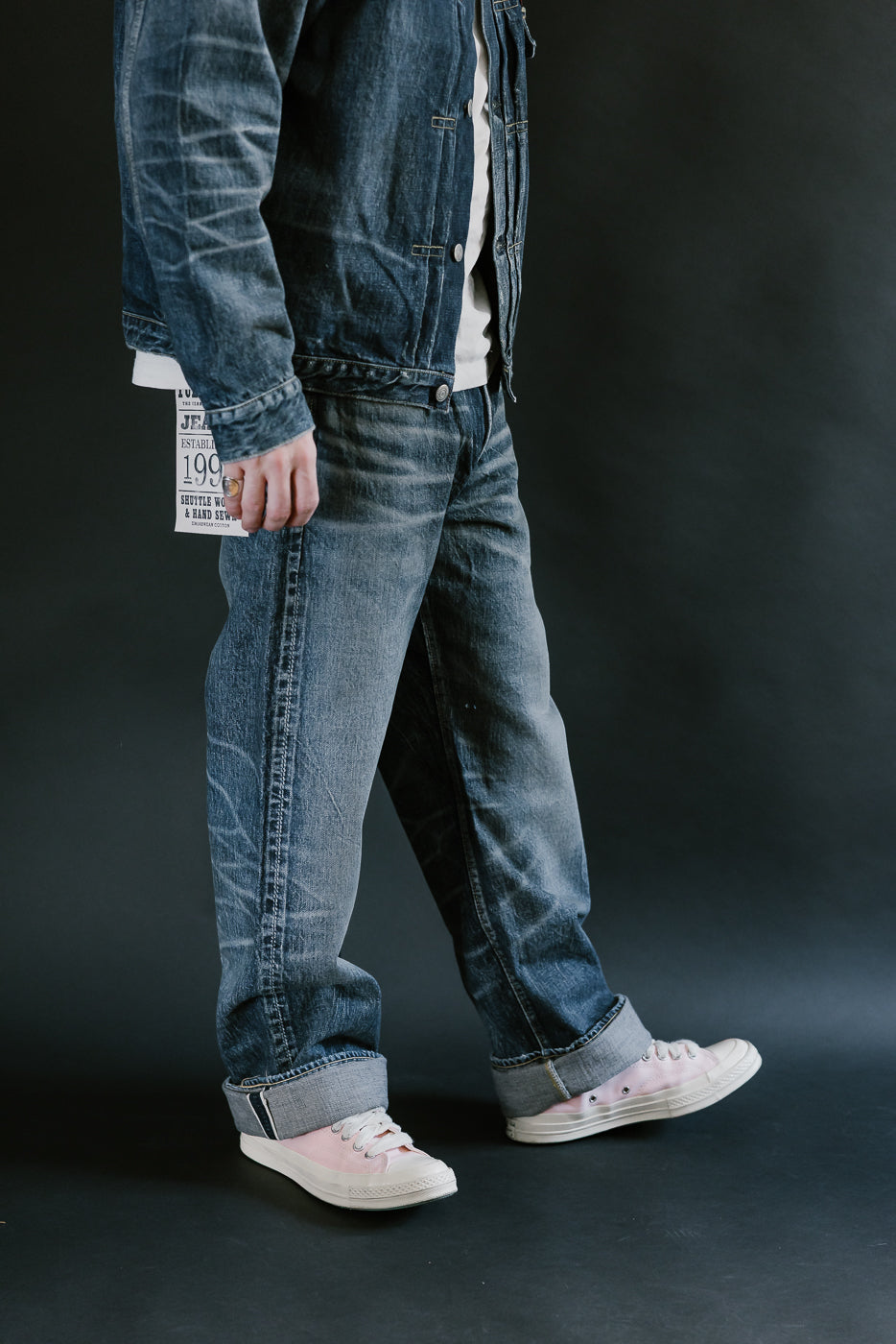 Man wearing heavily washed denim jacket and jeans,and pink shoes with a plain black background, 3/4 body, side view.